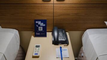 A hotel room with a bed, a bedside table with a phone, a pen, a notepad, and a sign with a QR code.