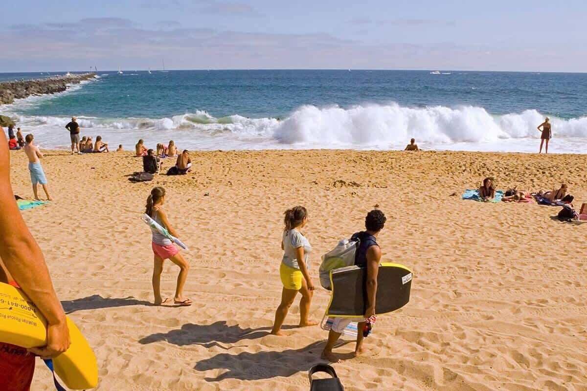 Una playa con gente caminando y sentada, olas rompiendo y barcos a lo lejos.
