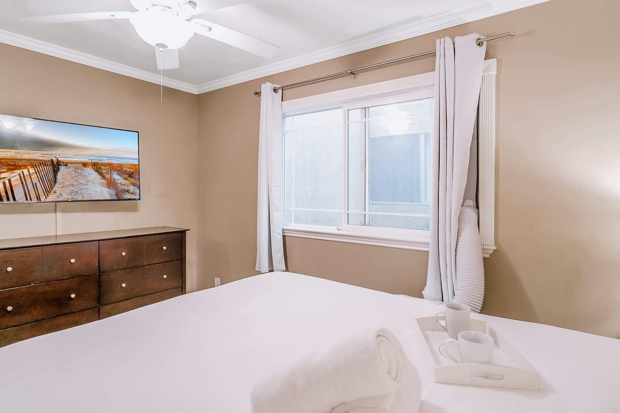 A bedroom with a bed, a TV on the wall, a window with curtains, and a ceiling fan.