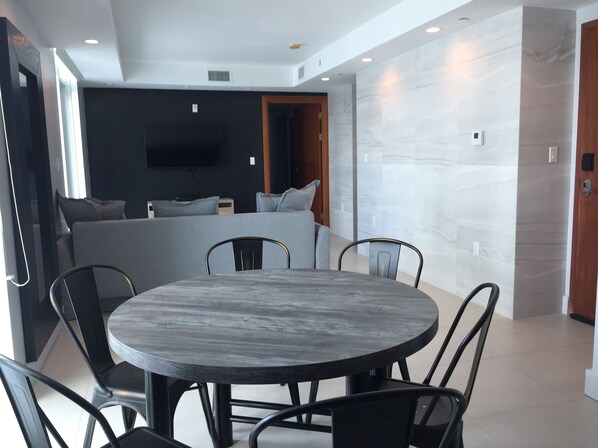 A modern living room with a round dining table, black metal chairs, a grey sofa, and a flat-screen TV mounted on the wall. - Miami Beach Luxury Condo, W/Private Terrace (301) (Miami Beach)