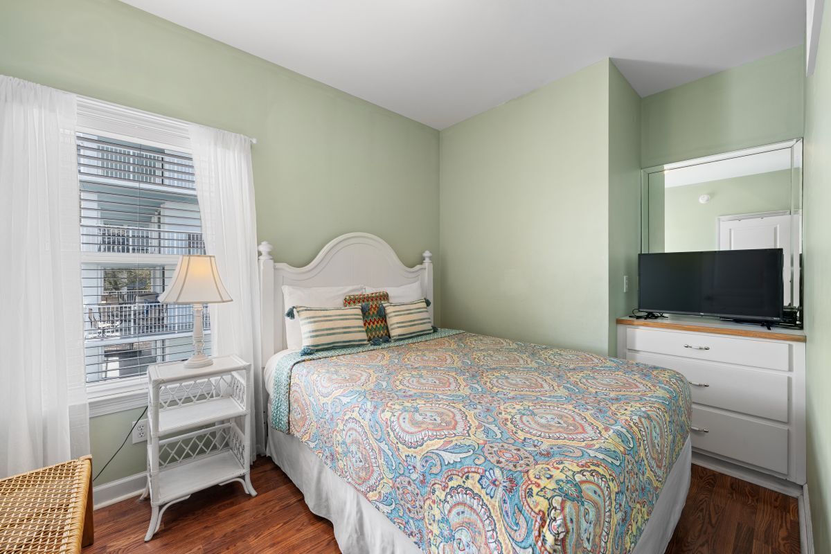 A bedroom with a bed, a TV, a window with blinds, and a bedside table.