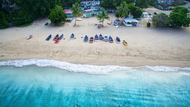 Vista aérea de una playa con varios barcos alineados en la arena y algunos edificios al fondo.