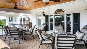 A covered patio with a wooden ceiling, a table with chairs, and a view of the house through large windows.
