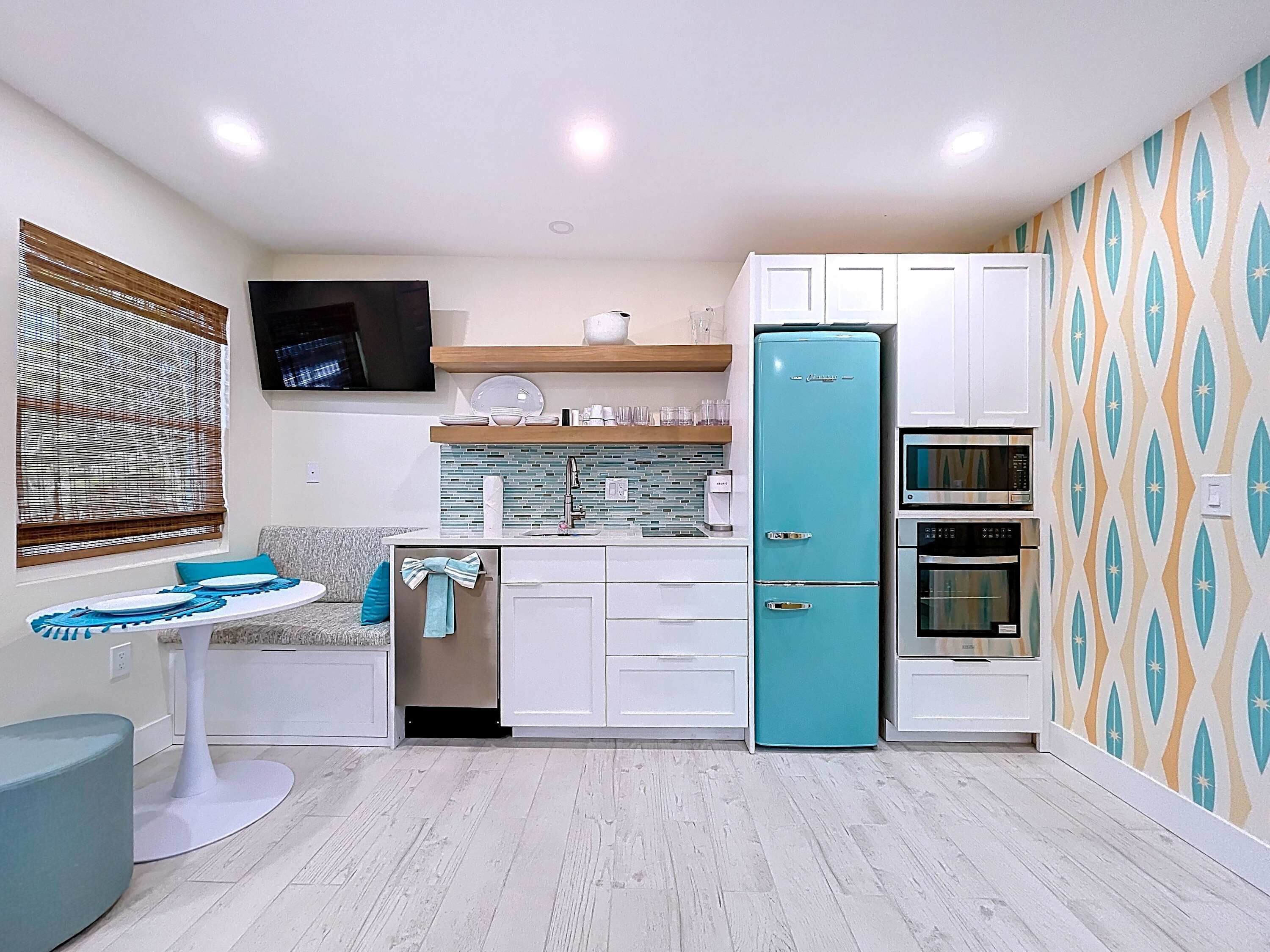 Een kleine keuken met een blauwgroene koelkast, witte keukenkastjes, een magnetron en een oven. Er staat een eettafel met blauwe placemats en een bank.