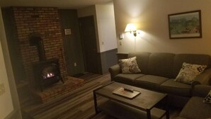 A living room with a brick fireplace, a sofa, a coffee table, and a wall-mounted lamp. - ☆ SKI ON & OFF ☆ Spruce Glen B on Great Eastern Trail w/AC, Fireplace, Sauna (Killington)