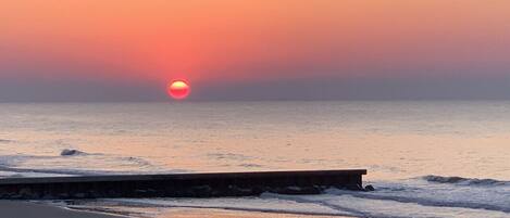 A beach scene with a sunset over the ocean, a wooden pier extending into the water, and a sandy shore.