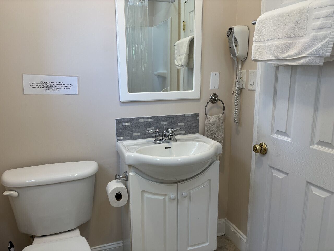 A bathroom with a toilet, sink, and a mirror. There is a sign on the wall and a hairdryer on the wall.