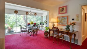 Una habitación amplia con un gran ventanal, un comedor con sillas y una mesa, un escritorio con lámpara, y un florero con flores.
