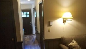 Un couloir avec un plancher en bois, un canapé agrémenté d’un coussin à motifs, une lampe fixée au mur et une porte au fond.