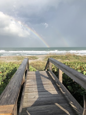 Une promenade en bois menant à une plage où l’on peut voir des vagues et un arc-en-ciel dans le ciel.
