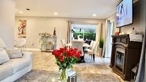 Een moderne woonkamer met een witte bank, een glazen salontafel met een vaas rode rozen, een open haard en een aan de muur bevestigde televisie.