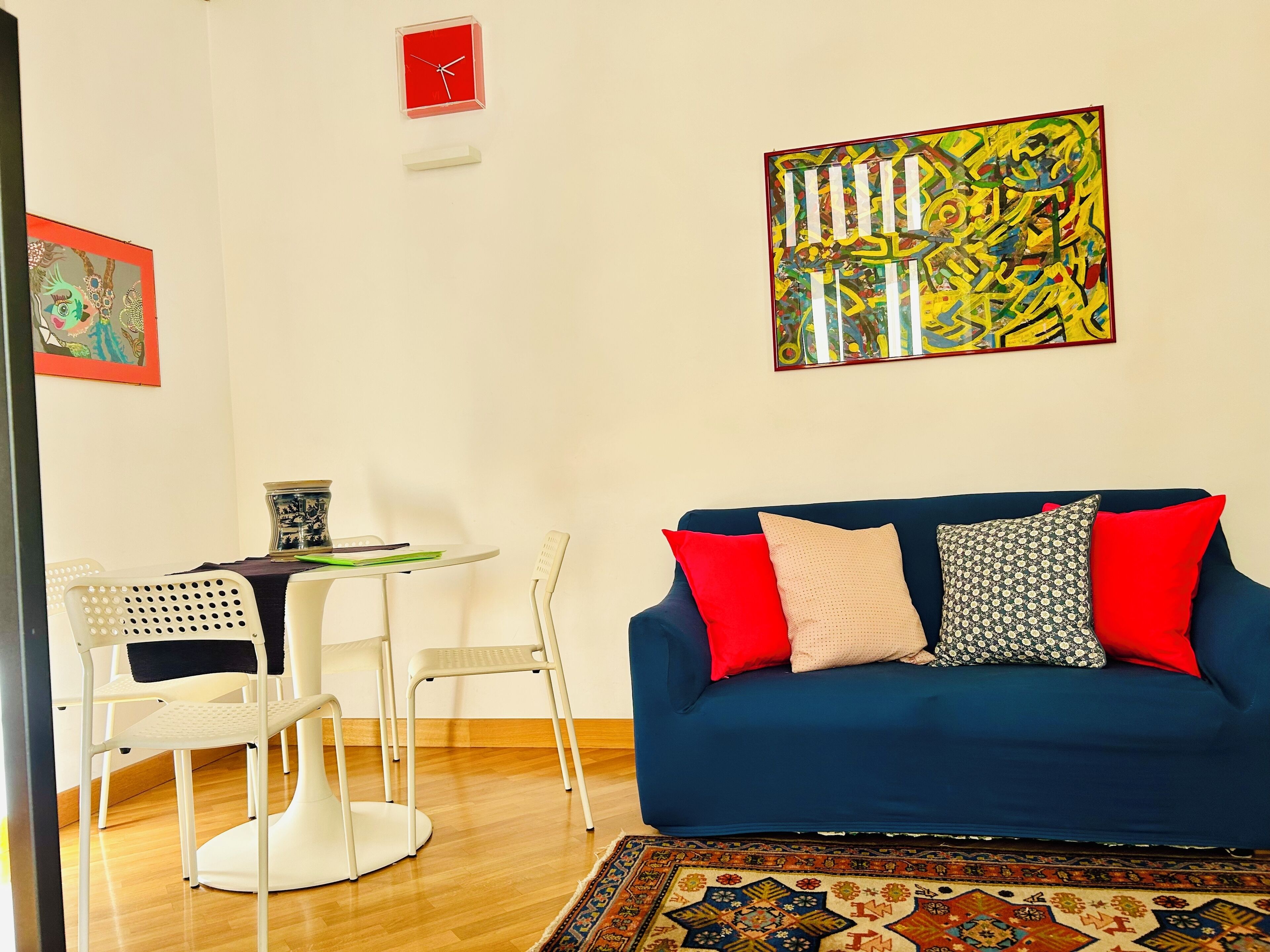 Un salon avec un canapé bleu, des chaises blanches, une petite table ornée d’un vase décoratif et un tableau abstrait aux couleurs vives accroché au mur.