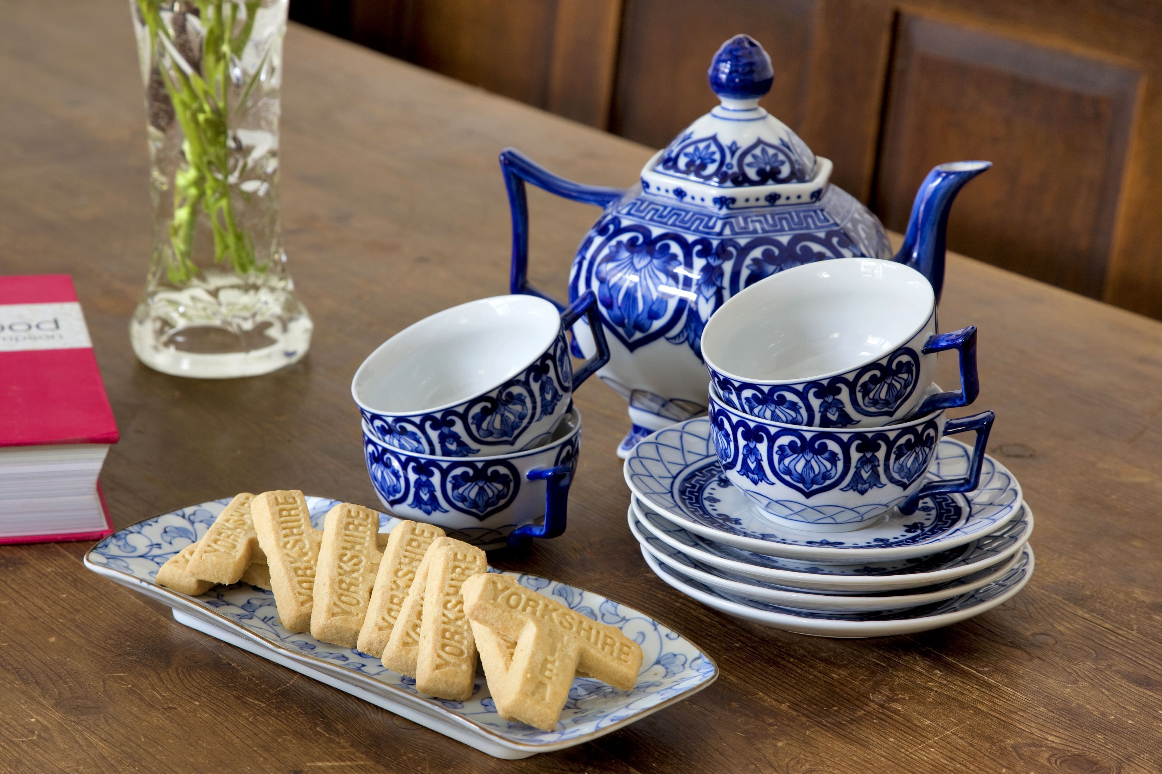 A table setting with blue and white porcelain tea set, a plate of Yorkshire biscuits, and a pink book.