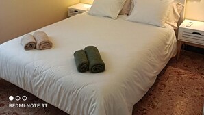 A hotel room with a neatly made bed, two rolled towels, a painting on the wall, and bedside lamps.