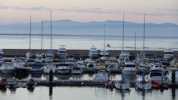 A marina with various boats docked, a calm water body, and distant mountains.