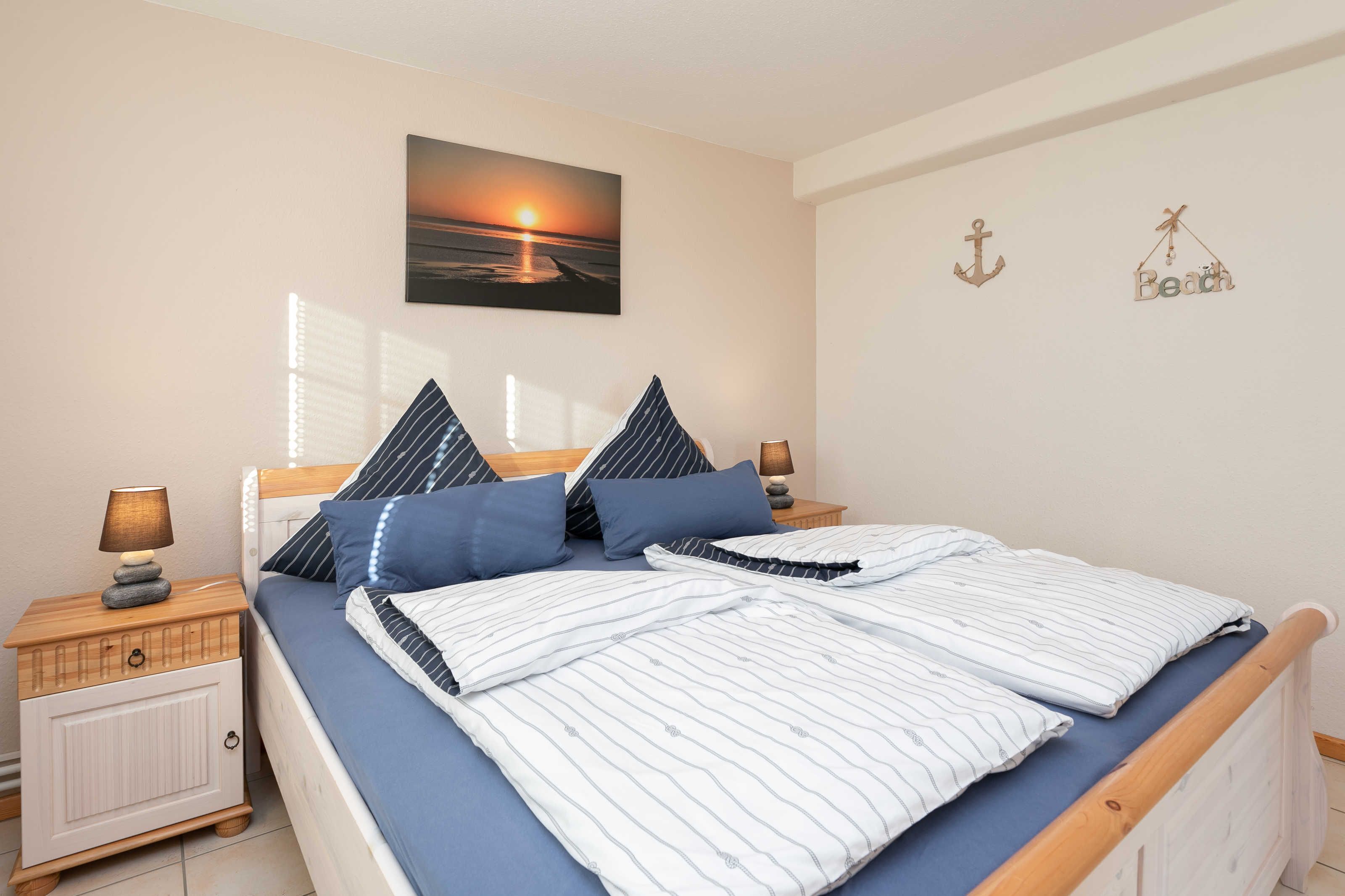 Een net opgemaakt bed met gestreepte lakens, een nachtkastje met een lamp en een aan de muur hangend schilderij van een zonsondergang.