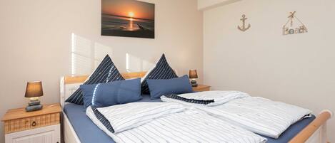 Een net opgemaakt bed met gestreepte lakens, een nachtkastje met een lamp en een aan de muur hangend schilderij van een zonsondergang.