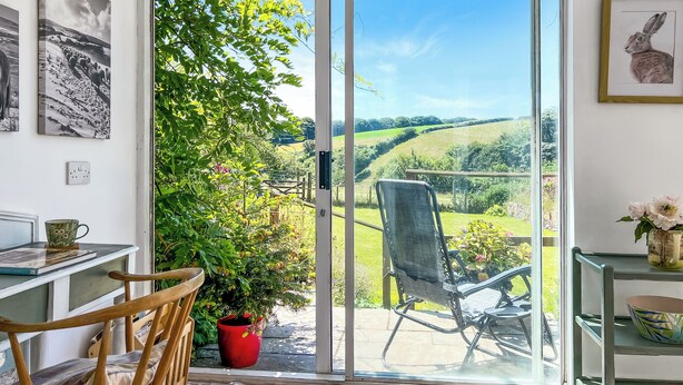 A room with a sliding glass door leading to a garden, a wooden chair, a table with a mug, and a shelf with a vase. - Brook Cottage, Challacombe (Challacombe)