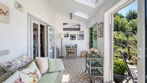 A sunlit room with a patterned floor, a sofa with floral cushions, a desk with a lamp, and a view of greenery outside. - Brook Cottage, Challacombe (Challacombe)