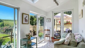 A bright room with a glass door leading to a garden, a sofa with cushions, a small table with a plate of food, and a glass shelf with items. - Brook Cottage, Challacombe (Challacombe)