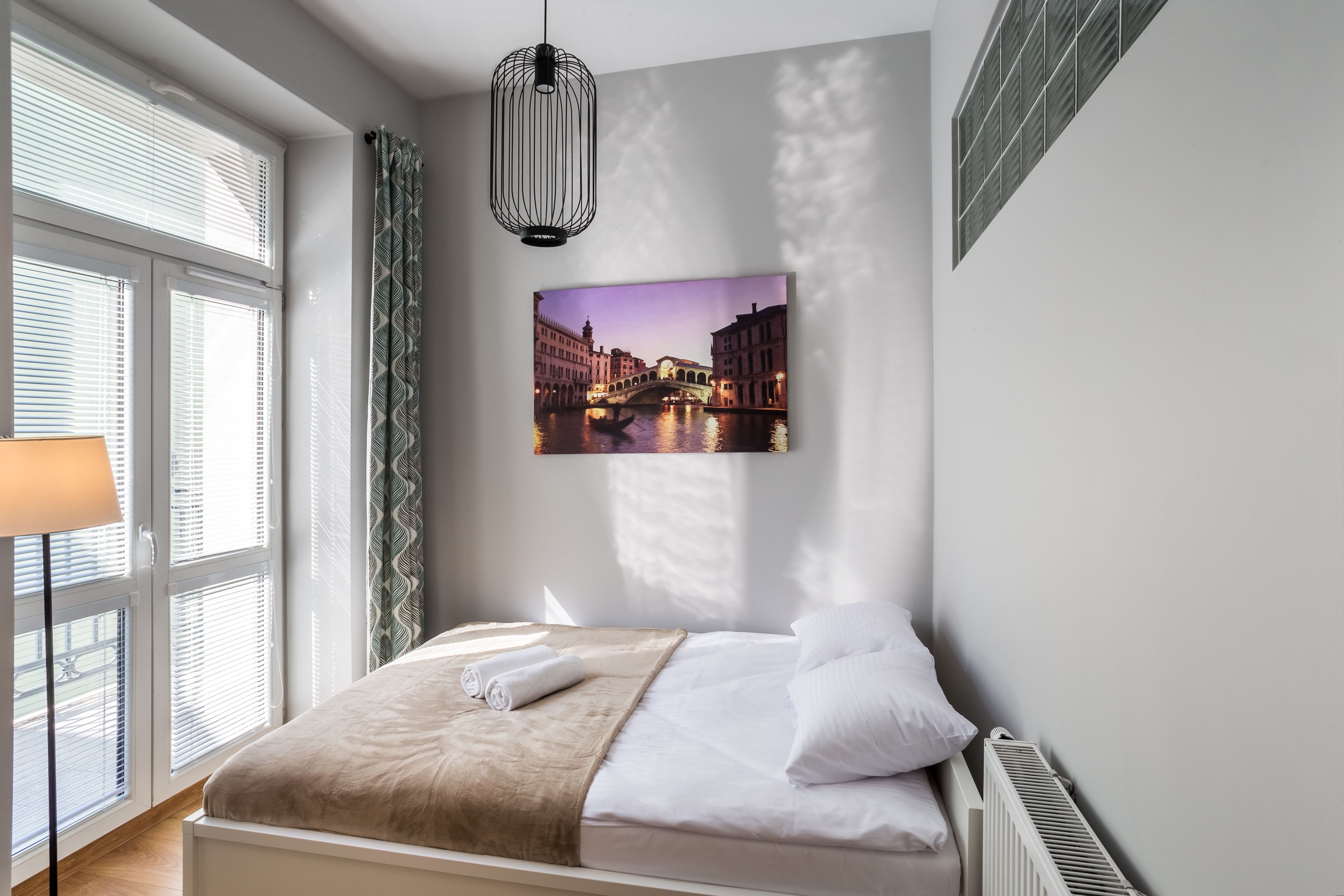 A neatly made bed with a blanket and pillows, a wall-mounted picture of a cityscape, a window with blinds, and a floor lamp.