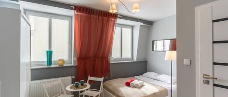 Un dormitorio pequeño con cama, una mesita, sillas, un radiador y una ventana con cortinas.