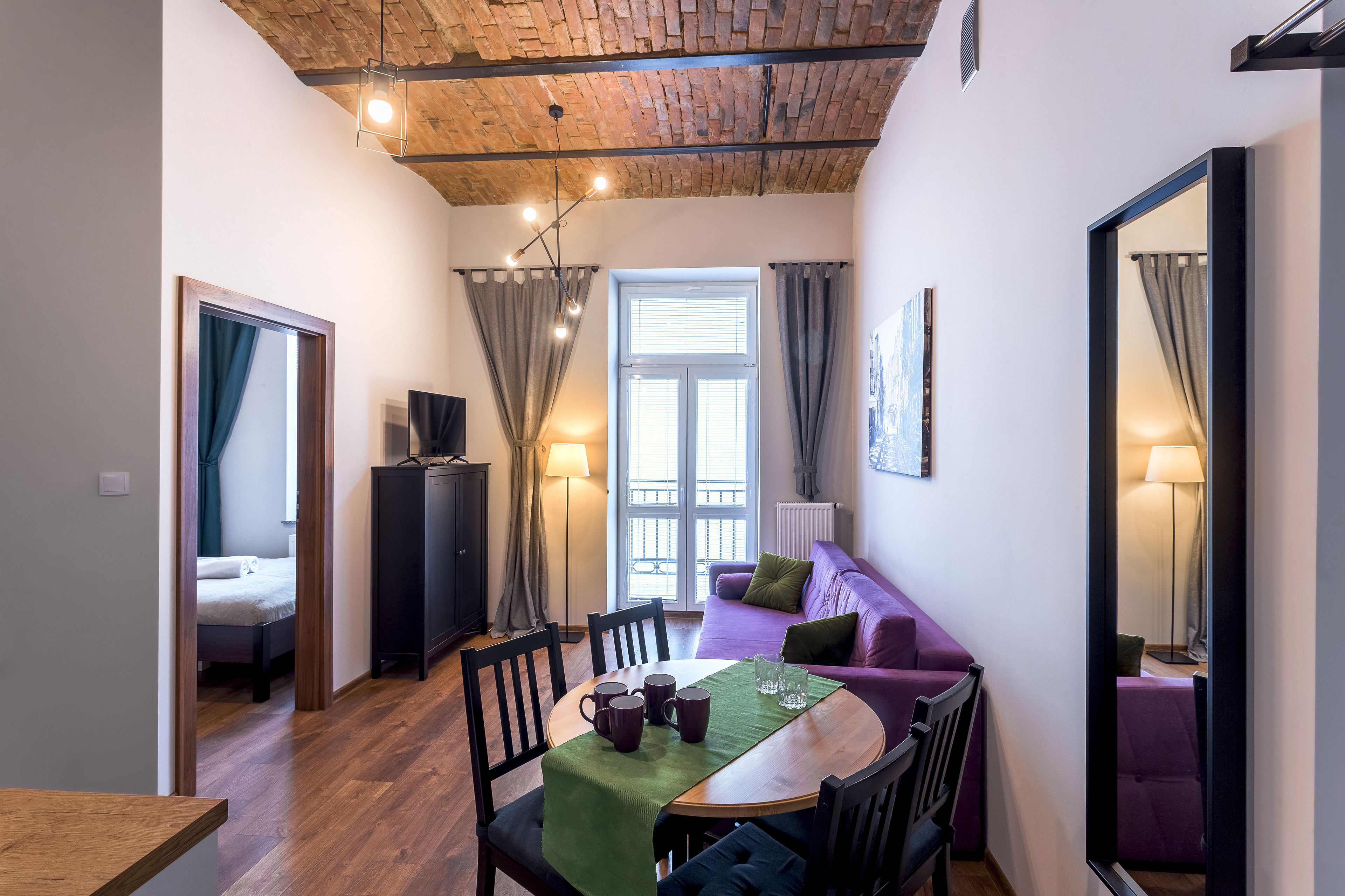 Una habitació acollidora amb sostre de fusta, una taula de dinar amb cadires, un sofà morat i una finestra gran amb cortines.