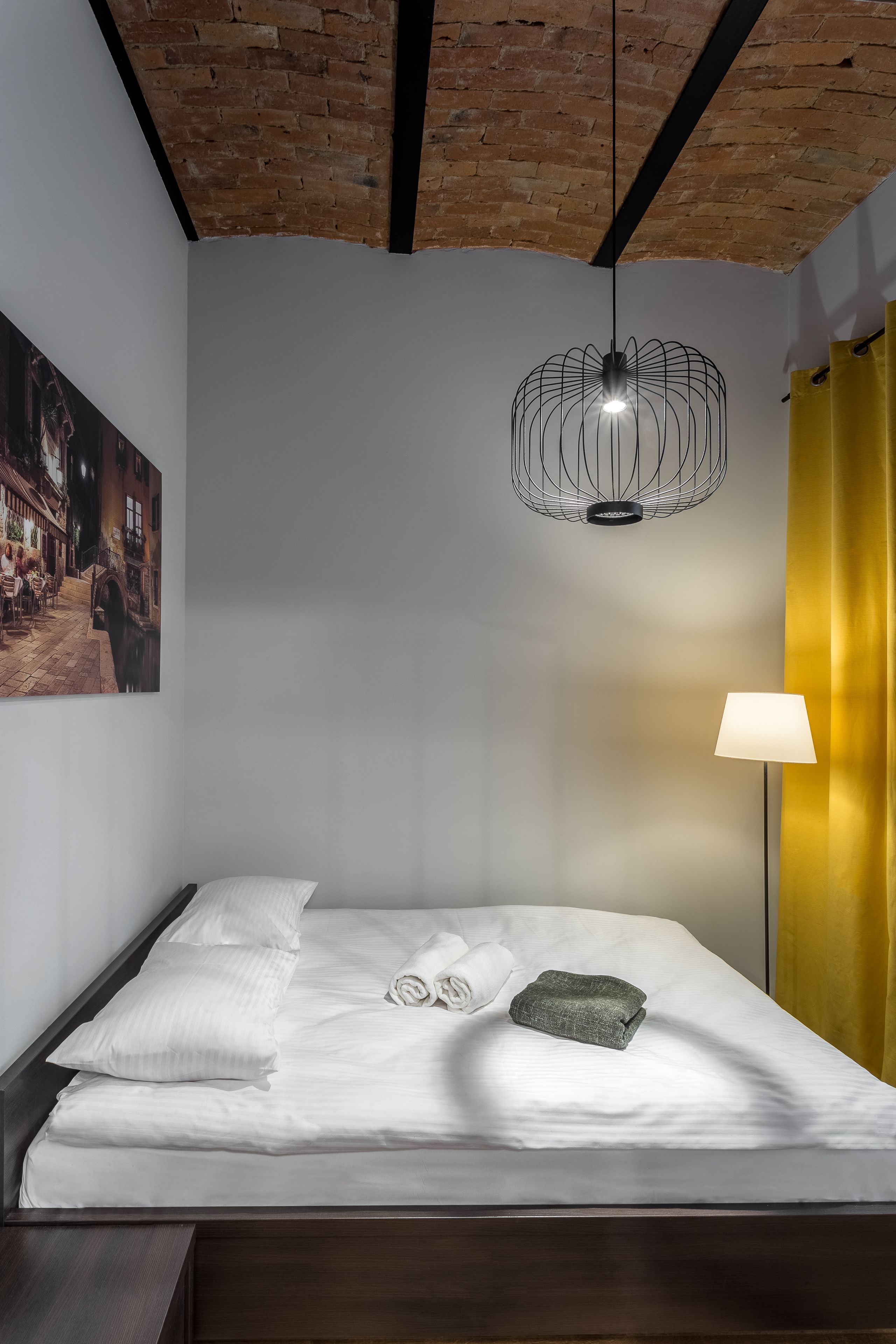 A neatly made bed with white linens, a green towel, and a rolled towel on a wooden headboard. A modern pendant light hangs above the bed, and a painting of a street scene is visible on the wall.