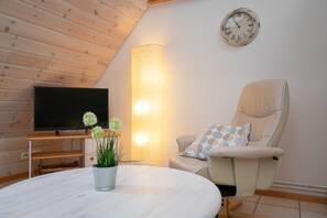 A cozy living room with a flat-screen TV, a comfortable armchair, a round table with a potted plant, and a wall-mounted clock.