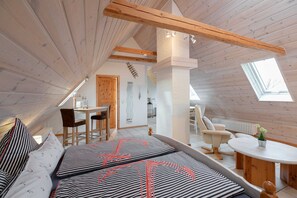 A cozy attic bedroom with a sloped ceiling, wooden beams, a skylight, a dining area, and a seating area with a table and chairs.