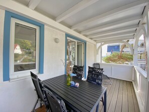 Uma área de estar coberta ao ar livre, com mesa e cadeiras, vista para uma casa através de uma janela e um corrimão.