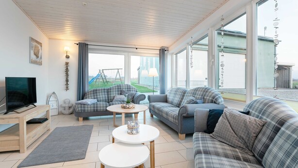 Un salon spacieux comprenant un grand canapé, un canapé plus petit, une table basse et un téléviseur posé sur un meuble en bois. De grandes fenêtres offrent une vue sur l’extérieur.