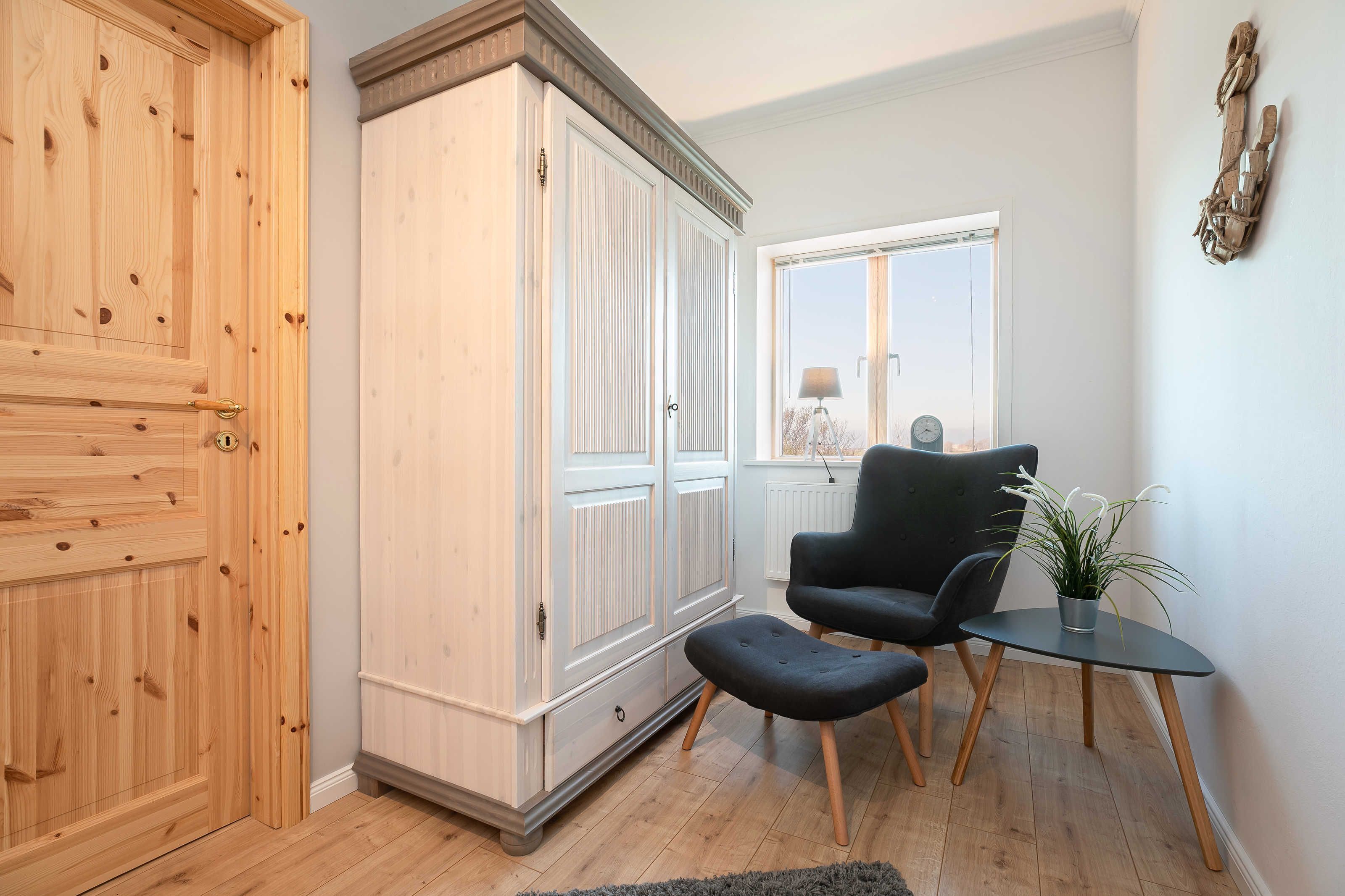 Una habitación con un armario de madera, un sillón negro, una mesita con una planta y una ventana que ofrece vistas al exterior.