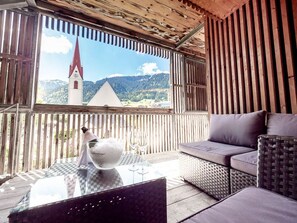 A wooden balcony with a view of a church and mountains, a glass table with a bottle and glasses, and wicker furniture.