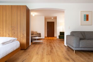 A room with a bed, a grey sofa, a wooden wardrobe, a small round table, and a door leading to another room. - Junior Suite - Hotel Krone Lingg GmbH & Co KG (Au)