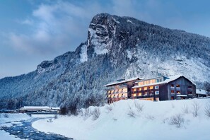 A mountain lodge with a river in front, surrounded by snow-covered trees. - Junior Suite - Hotel Krone Lingg GmbH & Co KG (Au)
