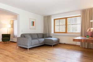A modern living room with a grey sectional sofa, a green armchair, a wooden floor, a window with blinds, and a radiator.