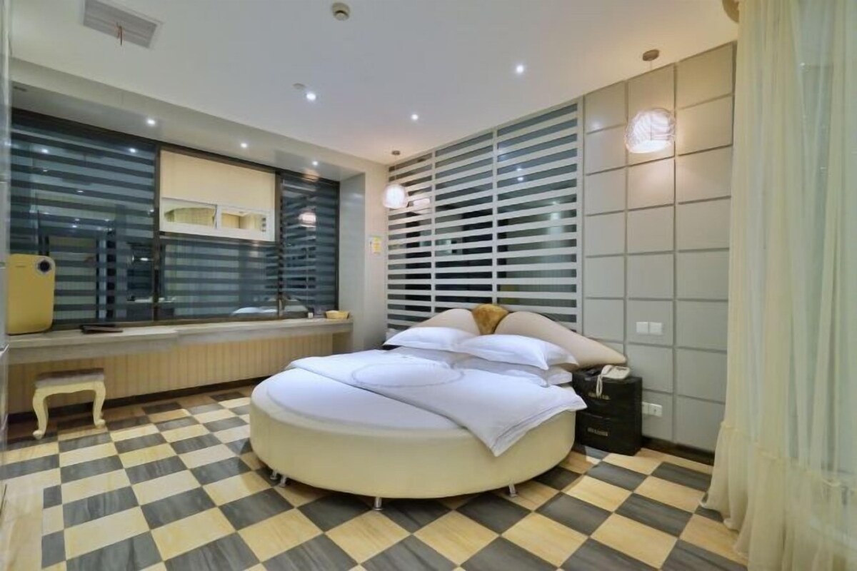 A modern bedroom with a round bed, a checkered floor, a window with blinds, and a bedside table.
