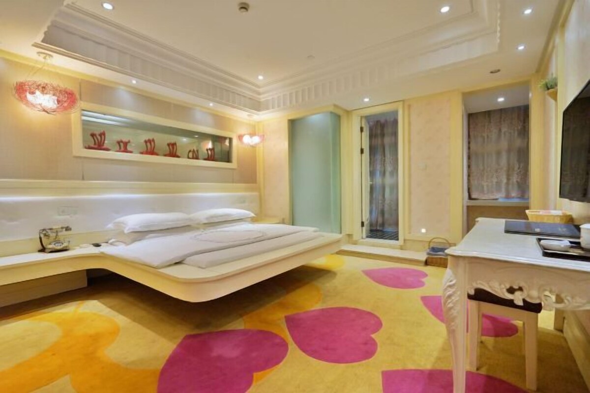 A modern hotel room with a curved bed, a bedside table, a small desk, and a large window with curtains.