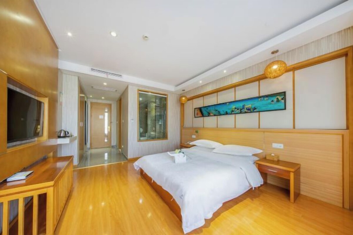 A hotel room with a wooden floor, a bed with white linens, a wooden nightstand, a television on a stand, and a painting on the wall.
