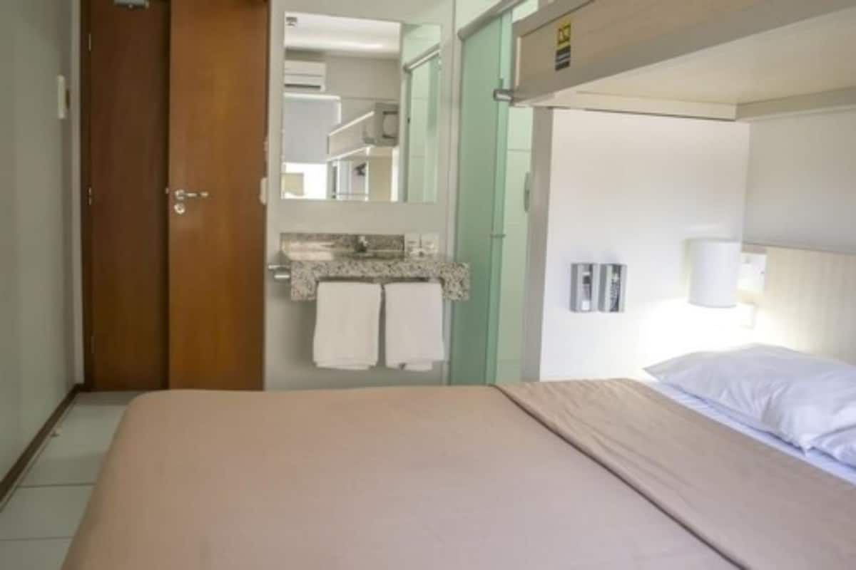 Habitació d'hotel amb un llit, un bany amb un lavabo i tovalloles, i una porta que condueix a una altra habitació.