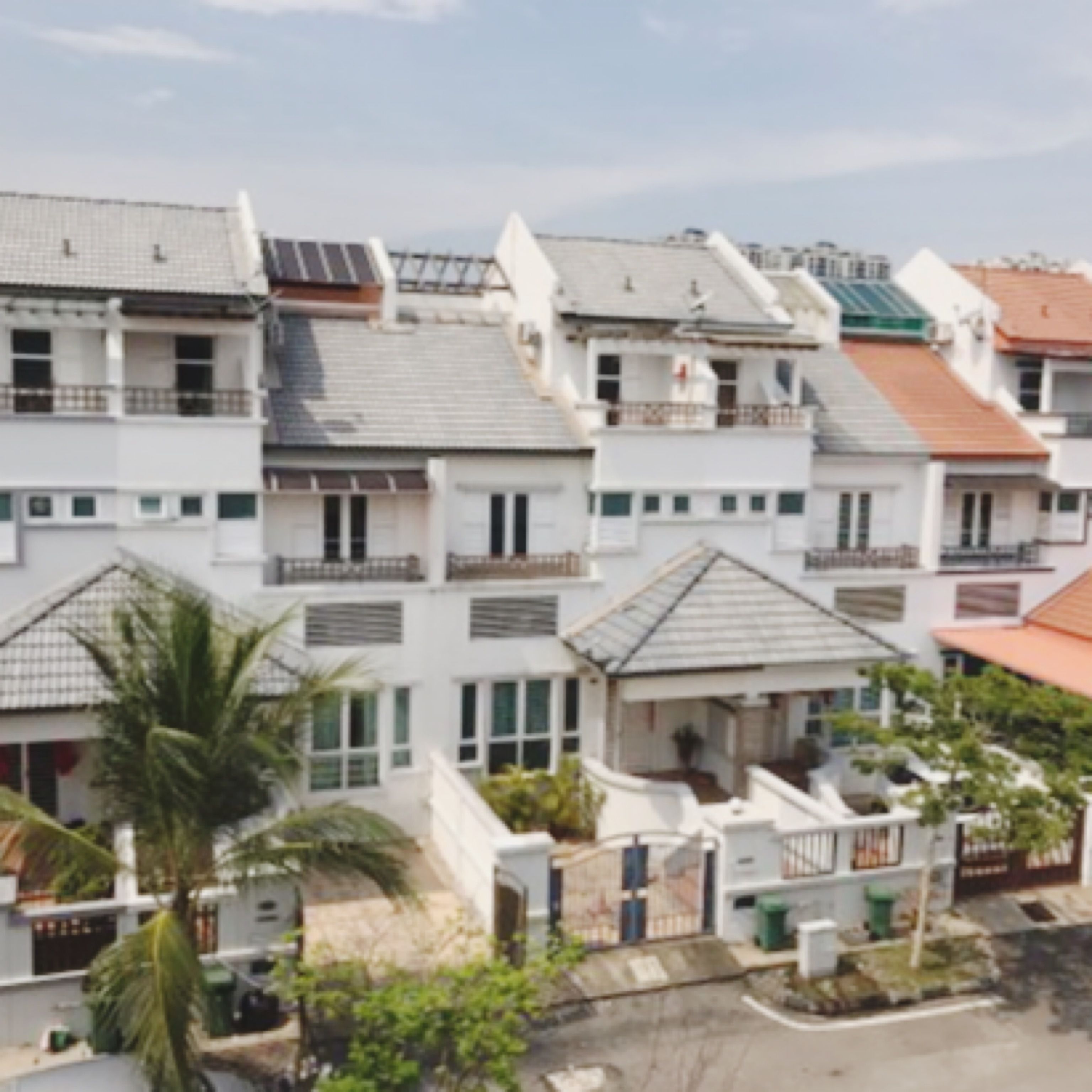 A row of white residential buildings with flat roofs, some with solar panels, and a gated entrance with a palm tree in the foreground.