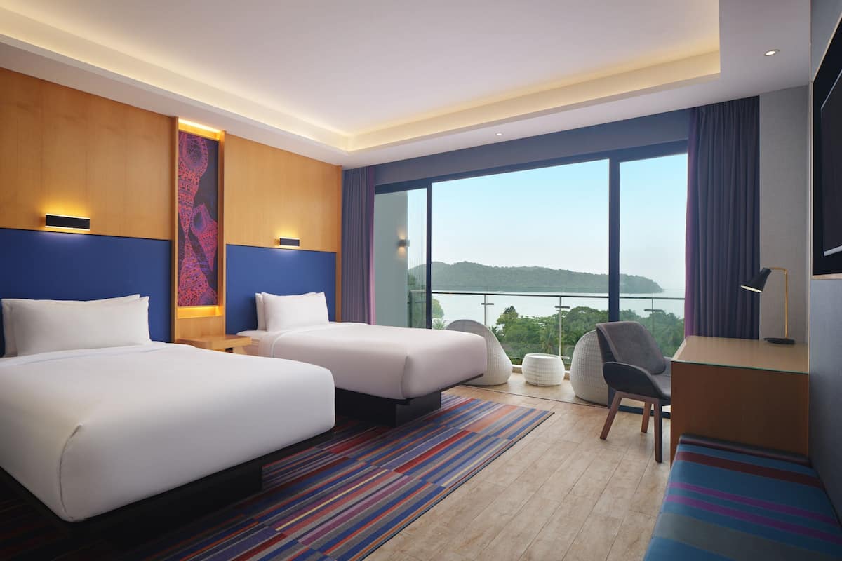 Une chambre d’hôtel avec deux lits, un bureau, une chaise, une grande fenêtre donnant sur un beau paysage et un tapis coloré.