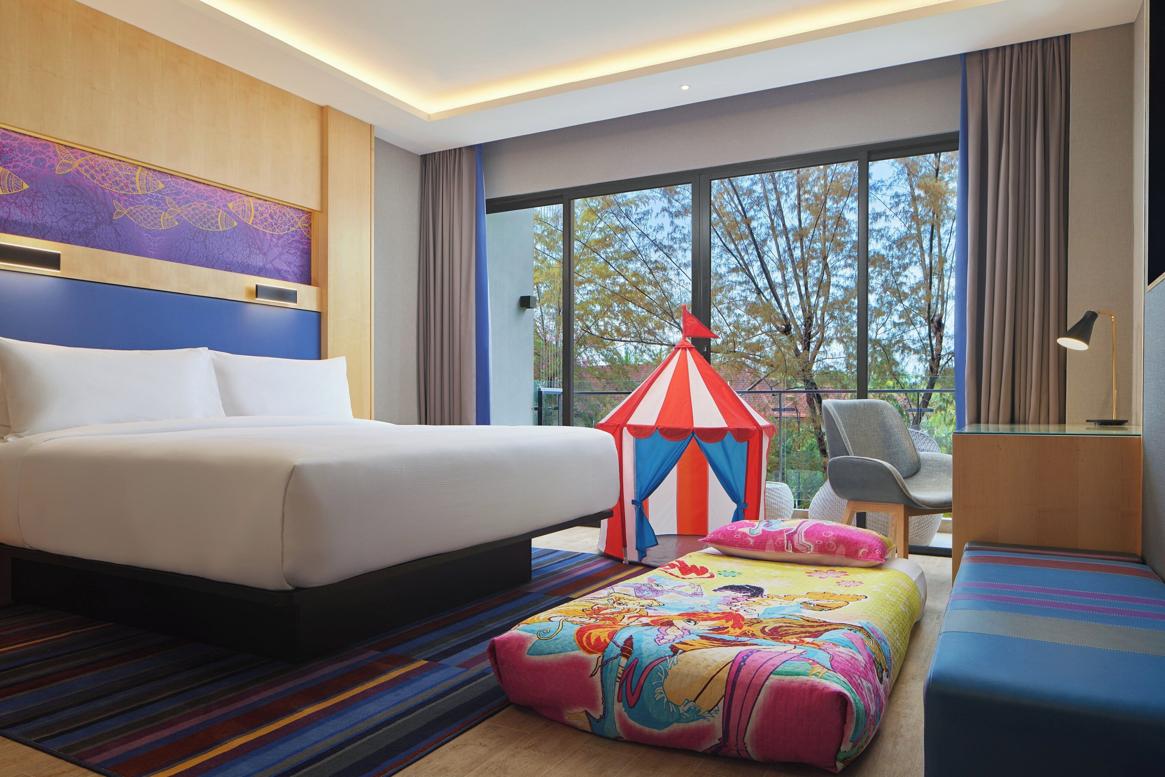 Une chambre d’hôtel avec un grand lit, un pouf coloré, une petite structure ressemblant à une tente, une chaise et une vue sur les arbres par la fenêtre.