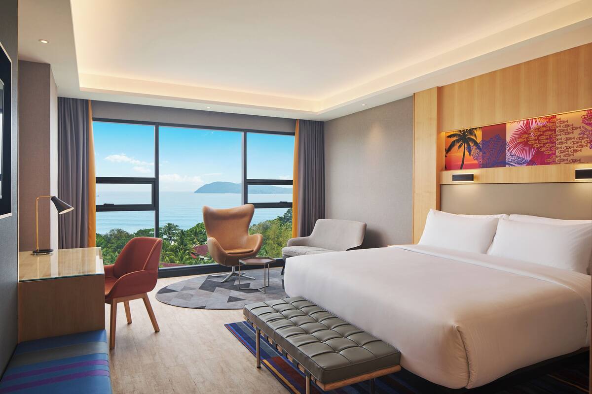 Une chambre d’hôtel avec un lit, un bureau, une chaise et une grande fenêtre donnant sur un beau paysage.