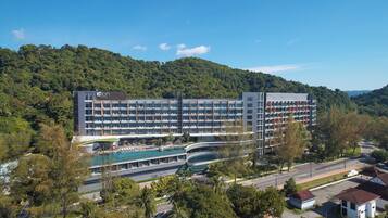 A multi-story hotel with a swimming pool, surrounded by lush greenery and a hilly backdrop.
