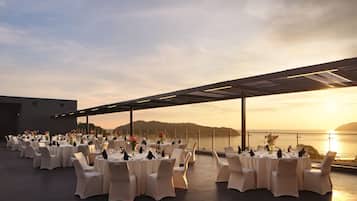 An outdoor dining area with round tables and white chairs, set against a sunset view.