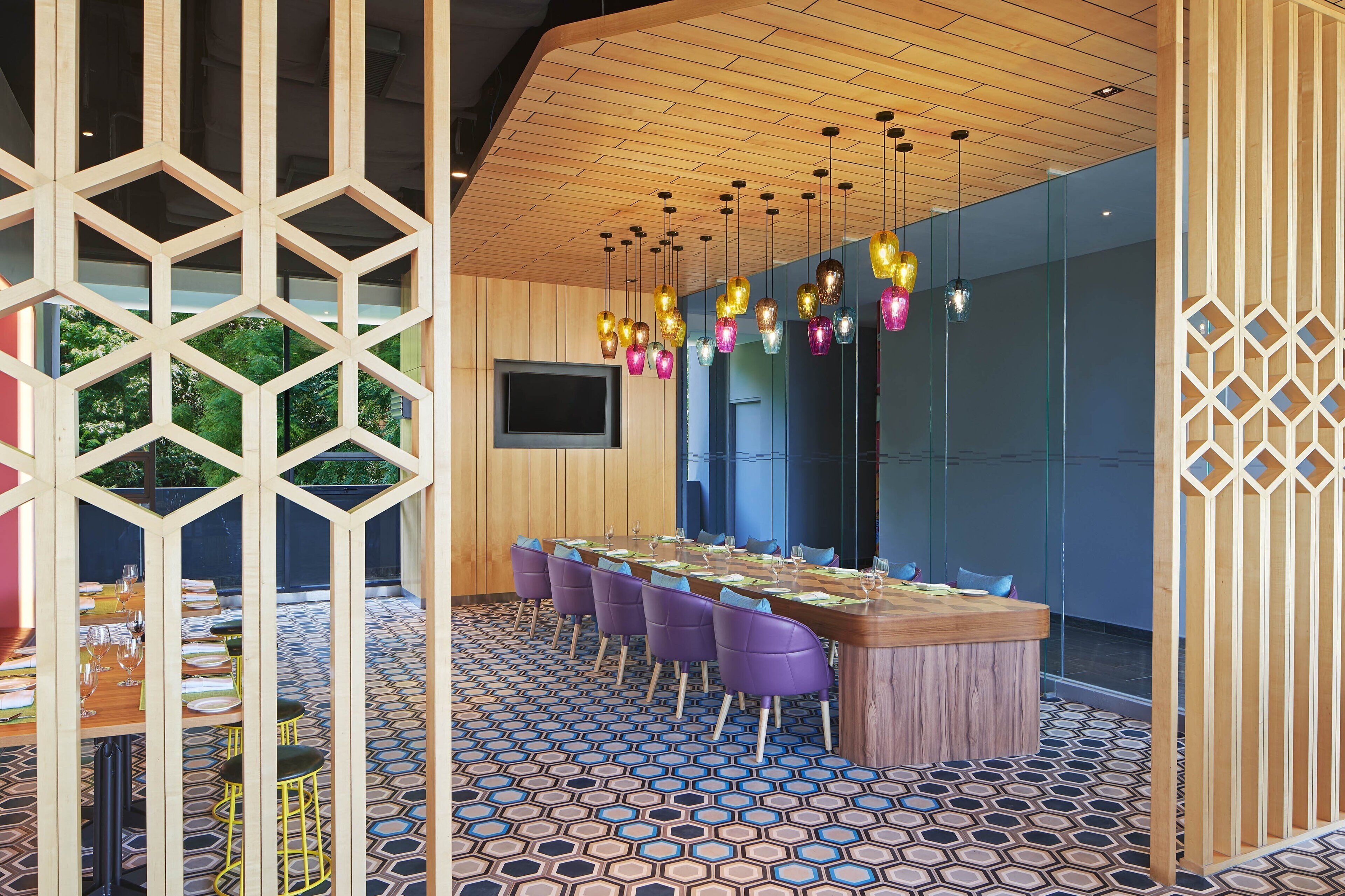 A modern dining area with a long wooden table, purple chairs, and colorful hanging lights.