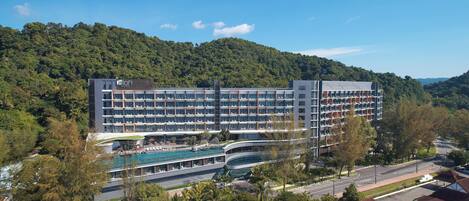 Un hotel moderno con varios pisos, una piscina y una exuberante colina verde al fondo.
