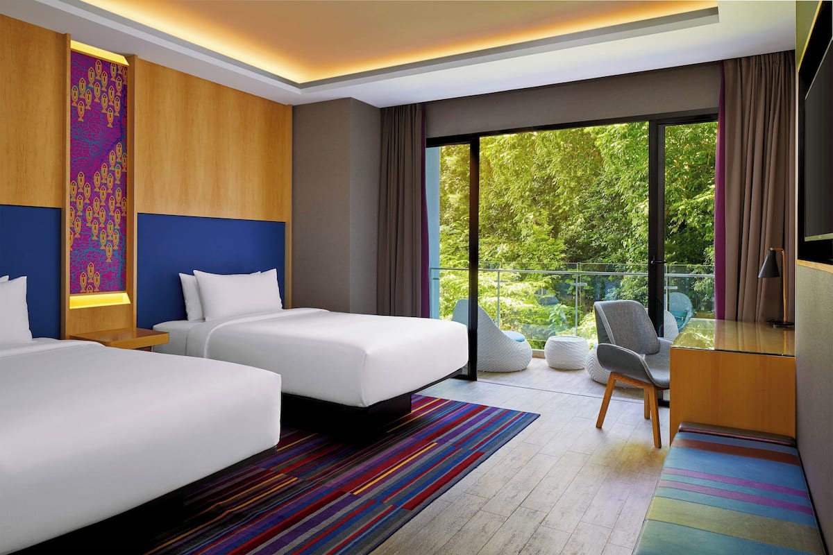 Une chambre d’hôtel moderne, équipée de deux lits, d’un bureau, d’une chaise et d’un balcon offrant une vue sur la verdure.